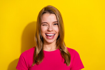 Smiling carefree young woman wearing pink t-shirt winking playfully against a bright yellow...