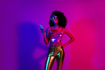 Trendy young woman in colorful neon lighting holding a phone and smiling confidently in a fashionable outfit