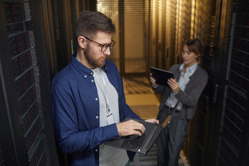 Two IT professionals managing server equipment in modern data center. One person is using laptop while another is using tablet, ensuring all systems are operational