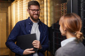 Smiling IT professional holding laptop while talking with colleague in server room surrounded by...
