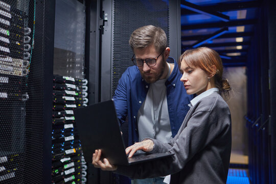 Two IT professionals are examining server connections in a data center, focusing on laptop screen while discussing necessary steps for troubleshooting issues - Powered by Adobe