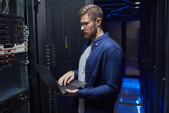 Bearded man in blue shirt engaged in managing servers in a modern data center, holding a laptop while examining server racks for maintenance and monitoring purposes