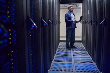Male technician standing in data center while using laptop for server performance monitoring and...