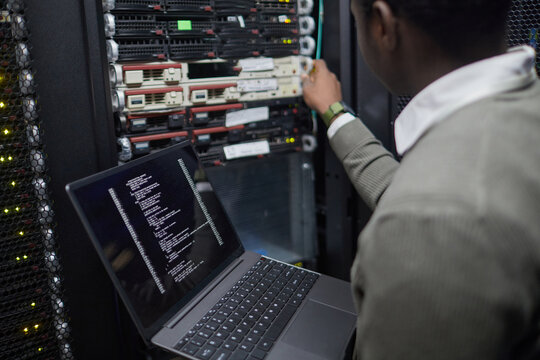 Person managing server equipment in a technology environment, checking system status on a laptop. Focus on information technology and network administration