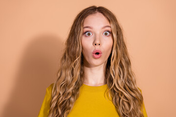 Surprised young woman with long wavy hair wearing yellow t-shirt against beige backdrop