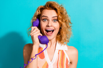 Naklejka premium Cheerful Young Woman With Red Hair Smiling Excitedly While Talking on a Retro Purple Phone Against a Vibrant Teal Background