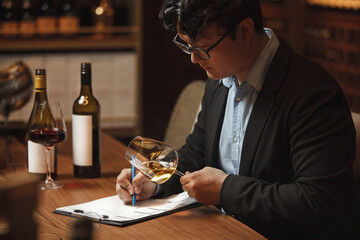 Wine Expert Evaluates Color of White Wine Over Clean Paper Background Sitting at Table in Cellar
