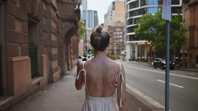 A woman in a light, backless dress walks along a city street in Sydney, surrounded by a mix of historic sandstone buildings and modern glass skyscrapers.
