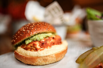 Crispy chicken burger topped with guacamole on a sesame seed bun in a warm, ambient restaurant setting, soft focus. Vertical image