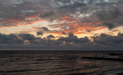 Silhouette of person standing on pier against sea at sunrise with dramatic clouds and orange sky. Vertical seascape photography. Travel and solitude concept for poster and wallpaper