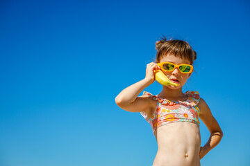 Little girl in sunglasses holding a banana like a phone under a bright blue sky, with copy-space