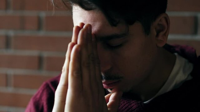 A young man sits with eyes closed and hands joined, praying quietly for inner peace and strength during a moment of emotional need