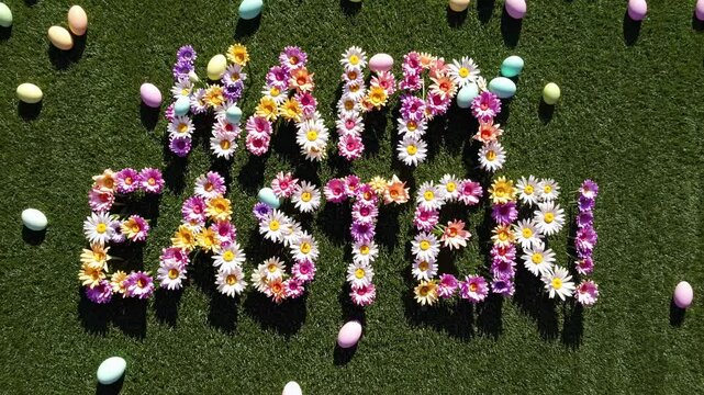 Top-down view of 'Happy Easter' spelled with flowers on grass, surrounded by colorful eggs. Bright, festive style ideal for a holiday video.