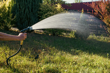 Male hand holding garden hose sprayer watering lawn in backyard on sunny day