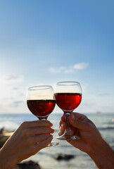 Male and female hands with glass goblets of red wine against the sky on the seashore. Romantic evening for two in nature. Valentine's Day celebration