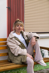 Young stylish woman sits on park bench with one leg carelessly resting on seat, exuding ease and...