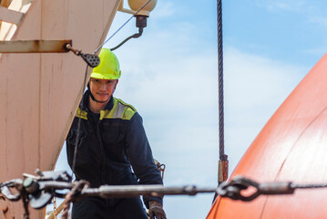 Portrait of the young Filipino officer, fully focused on his task during the inspection of the lifeboat and rescue boat. Dressed in a blue coverall and yellow helmet, he works with precision and care.