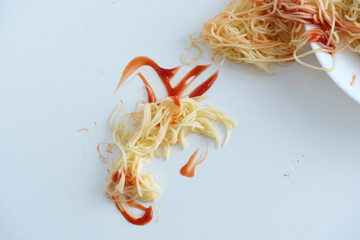 A plate of noodles partially toppled over a clean white surface, with ketchup drizzled amongst the...