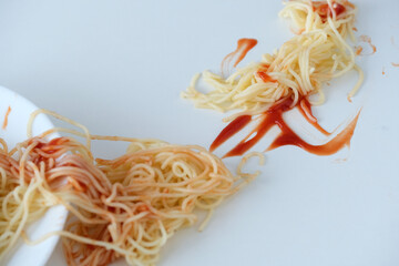A plate of noodles partially toppled over a clean white surface, with ketchup drizzled amongst the...