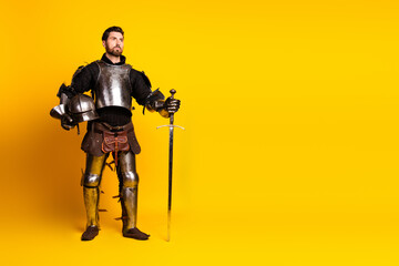 Fototapeta premium Man dressed as a medieval knight in full armor holding a sword and helmet against a vivid yellow background representing historical themes.