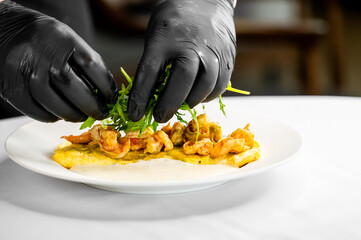 A chef in black gloves carefully garnishes a delicious omelet topped with shrimp and fresh arugula on a white plate, showcasing culinary skill and attention to detail in plating.