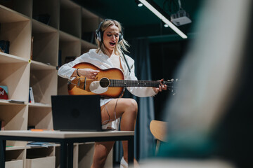 A woman wearing headphones sings and plays the guitar enthusiastically in a modern studio setup.