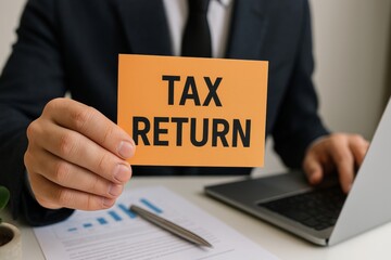 Obraz premium Businessman is holding an orange card with the words tax return written on it, while working on a laptop with financial documents on his desk