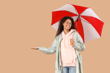 Happy young African-American woman in raincoat with umbrella on beige background