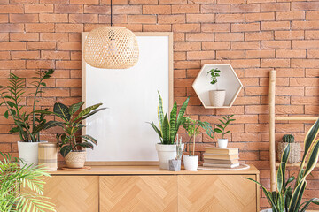 Green plants with books and blank frame on commode near brick wall in room