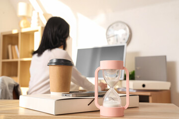 Hourglass with coffee cup, folder and mobile phone on table against businesswoman working in office, closeup