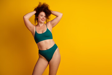 Obraz premium Smiling young woman with curly hair in green lingerie on yellow background embracing body positivity and confidence