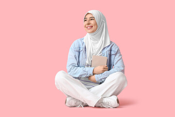 Beautiful young Muslim woman in hijab with book sitting on pink background