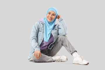 Beautiful young Muslim woman in hijab sitting on grey background