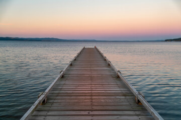 Fototapeta premium Empty wooden pier stretching into a calm lake at dusk, pastel pink and orange sky, tranquil scenery