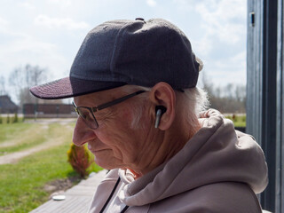 Senior man wearing baseball cap and hoodie enjoying music through wireless earbuds, standing against dark wooden background, experiencing peaceful moment of personal audio connection