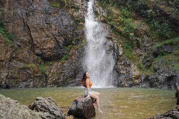 woman sits on rock by serene waterfall, surrounded by lush greenery and rocky cliffs