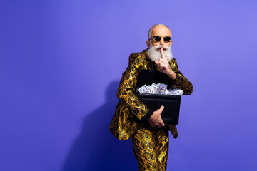 Stylish elderly gentleman with a white beard in a golden suit holding a briefcase full of cash showing a secret gesture
