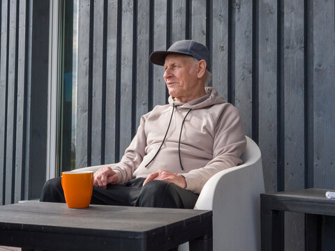 Comfortable elderly man wearing casual clothes and a cap sits in a modern white chair at a table. Savoring a warm beverage from an orange mug in a serene outdoor setting with a wooden wall background