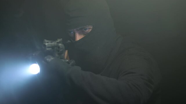 Burglar with black balaclava in the dark with gun and flashlight in hand. Strobe light and smoke. Burglar who broke into someone else's house.