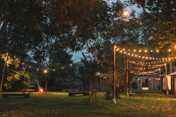 Cozy outdoor campfire surrounded by rustic wooden benches and glowing string lights, creating a...