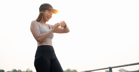 Asian woman checks her smartwatch while outdoors, dressed in activewear, enjoying a sunny day, embodying fitness and health. 