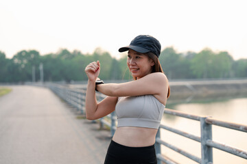 An asian woman stretches by a serene waterway, wearing a sports outfit and cap, emphasizing fitness...