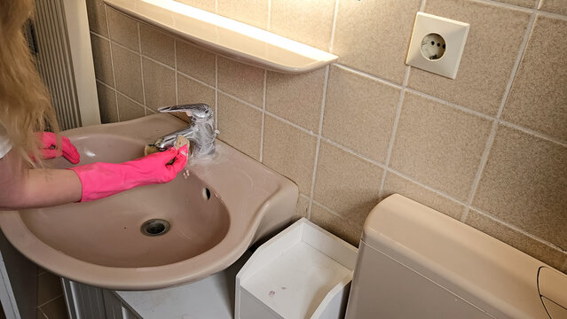 Person wearing pink gloves is cleaning a bathroom sink faucet with a scrub. Demonstrates household cleaning and hygiene practices. Emphasizes cleanliness and sanitation