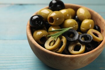 Different delicious marinated olive rings and rosemary in bowl on light blue wooden table, closeup. Space for text