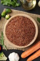 Raw brown rice in bowl among fresh products on dark textured table, flat lay