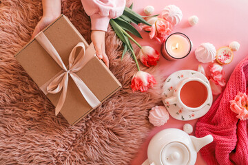 Top view of cozy feminine flat lay with hands holding a kraft gift box, pink tulips, cup of tea, candle, sweets, and a teapot on pink background with fur rug