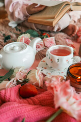 Romantic tea setup with a white teapot, pink tea in a floral cup, candle, roses, tulips, strawberries, and meringues arranged on a pink background with a cozy knitted fabric