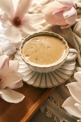 Elegant cup of cappuccino on a white saucer with blooming magnolia flowers on a wooden tray in soft morning light