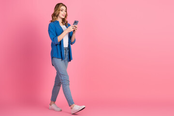 Young woman with blonde hair in casual blue shirt using smartphone against a pink background while...