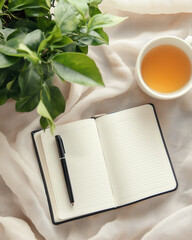 open notebook with pen rests on soft fabric beside cup of tea and lush green plant, creating serene and inviting atmosphere for journaling